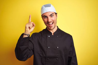 Üniformalı ve şapkalı genç ve yakışıklı aşçı izole edilmiş sarı arka planda başarılı bir fikri işaret ediyor. Heyecanlı ve mutlu. Bir numara..