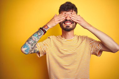 Young man with tattoo wearing striped t-shirt standing over isolated yellow background covering eyes with hands smiling cheerful and funny. Blind concept.