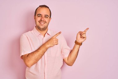 Zarif gömlekli genç adam pembe arka planda duruyor gülümsüyor ve kameraya bakıyor iki eliyle ve parmaklarıyla kenarı işaret ediyor..