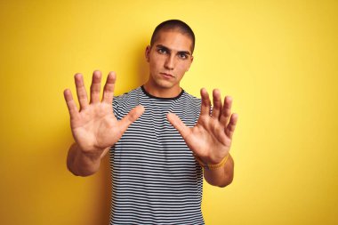 Sarı arka plan üzerinde çizgili tişört giyen genç yakışıklı adam korku ifadesinden korkuyor ve dehşete düşüyor. El hareketlerini durdur, şok içinde bağır. Panik konsepti.