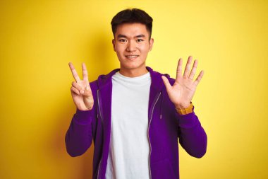 Mor kazak giyen Asyalı genç adam izole edilmiş sarı arka planda duruyor ve yedinci parmaklarını gösterirken kendinden emin ve mutlu gülümsüyor..