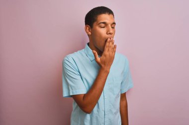 İzole edilmiş pembe arka planda duran mavi tişörtlü genç, yakışıklı Arap adam ağzıyla esnemekten sıkılmış. Huzursuz ve uykulu..