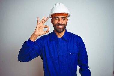 Üniforma ve kask takan yakışıklı Hintli işçi izole edilmiş beyaz arka planda gülümsüyor ve el ve parmaklarıyla tamam işareti yapıyor. Başarılı ifade.