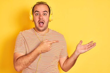 İzole edilmiş sarı arka planda duran kulaklıkla müzik dinleyen genç adam kameraya hayret ediyor ve gülümsüyor..