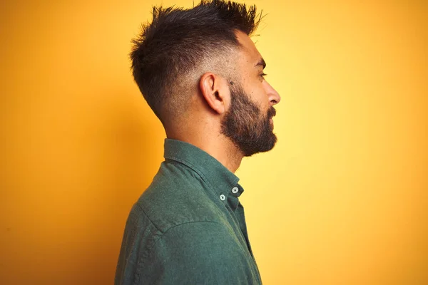 Young indian man wearing green shirt standing over isolated yellow background looking to side, relax profile pose with natural face with confident smile.