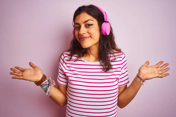 隔離されたピンクの背景にヘッドフォンを使用して音楽を聴いている若い美しい女性は 腕や手で提起された混乱した表現 疑いの概念 — ストック写真