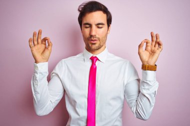 Tişört ve kravat takan genç yakışıklı işadamı pembe arka planda dikiliyor rahatlıyor ve parmaklarıyla meditasyon yaparken gözleri kapalı gülümsüyor. Yoga konsepti.