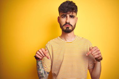 İzole edilmiş sarı arka plan üzerinde duran çizgili tişörtlü genç adam üzgün ve üzgün görünüyor, parmaklarıyla yönü gösteriyor, mutsuz ve depresif..