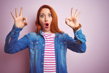 Güzel kızıl saçlı kadın kot gömlek giyiyor ve pembe arka planda çizgili tişört giyiyor. Şaşırmış ve şaşırmış görünüyor. Tamam sembolünü parmaklarıyla yapıyor. Çılgın ifade