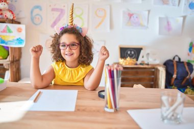 Gözlüklü ve tek boynuzlu at diademi takan güzel çocuk anaokulunda masanın üzerinde oturmuş kollarını kaldırarak ve açık gözlerle başarının şaşkınlığını ve şaşkınlığını kutluyor. Kazanan konsept.