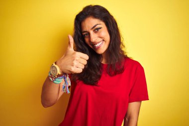 Kırmızı tişört giyen genç ve güzel bir kadın izole edilmiş sarı arka planda mutlu bir şekilde dikiliyor. Gülümsüyor.