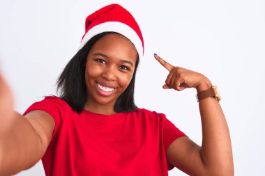 Genç Afrikalı Amerikalı kadın Noel şapkası takıyor ve izole bir arka planda selfie çekiyor. El ve parmağıyla işaret ederek çok mutlu.
