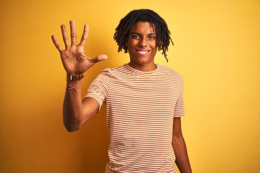 Rastalı tişörtlü Afro adam izole edilmiş sarı arka planda duruyor ve beş numarayı gösterirken kendine güvenen ve mutlu bir şekilde gülümsüyor..