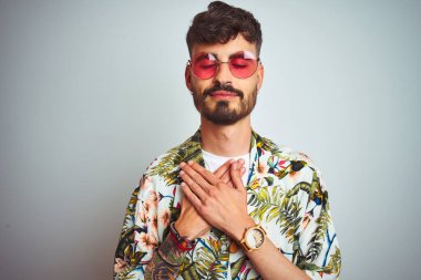 Tatilde dövmeli, güneş gözlüğü takan, izole edilmiş beyaz arka planda gülümseyen elleri göğsünde, gözleri kapalı ve yüzünde minnet ifadesi olan bir adam. Sağlık kavramı.