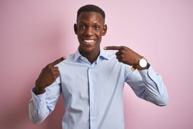 İzole edilmiş pembe arka planda duran mavi şık gömlekli Afro-Amerikan bir adam gülümseyen ve parmaklarıyla ve ağzıyla işaret eden. Diş sağlığı konsepti.