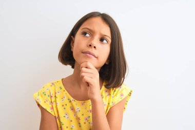 Sarı çiçekli elbise giyen güzel bir kız çocuğu izole edilmiş beyaz arka planda dikiliyor. Ciddi bir yüz ifadesi var. Soru sormayı düşünüyor.