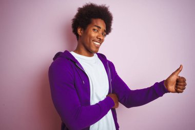 Mor kazak giyen genç Afro-Amerikalı adam pembe arka planda duruyor. Gururlu görünüyor, gülümsüyor. Kenarda baş parmak hareketi yapıyor.