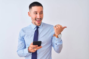 Genç yakışıklı iş adamı, izole edilmiş arka planda akıllı telefon kullanıyor ve gülümseyen bir gülümsemeyle baş parmağıyla onu işaret ediyor.