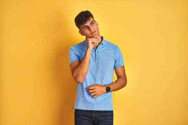 Mavi polo giyen genç Hintli adam izole edilmiş sarı arka planda duruyor ve elleri çenesinde soru sormayı, düşünceli ifadeyi düşünüyor. Düşünceli bir yüzle gülümsüyor. Şüphe kavramı.
