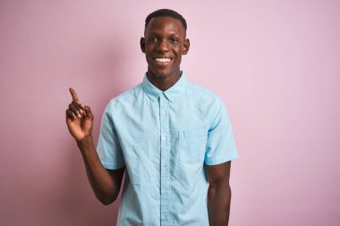 Mavi günlük gömlekli Afro-Amerikalı bir adam pembe arka planda duruyor yüzünde kocaman bir gülümseme, eli ve parmağı kameraya bakıyor..