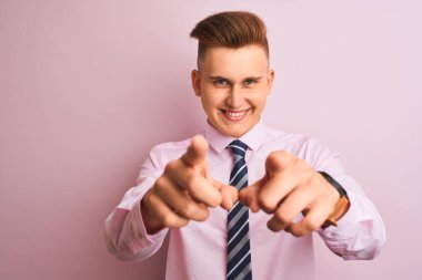 Genç yakışıklı iş adamı gömleği ve kravatı ile soyutlanmış pembe arka planda dikiliyor. Seni ve kamerayı parmaklarıyla gösteriyor.