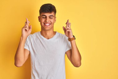 İzole edilmiş sarı arka planda duran beyaz tişörtlü genç Hintli adam umut ve gözler kapalı bir şekilde gülümseyerek işaret ediyor. Şans ve batıl inanç..