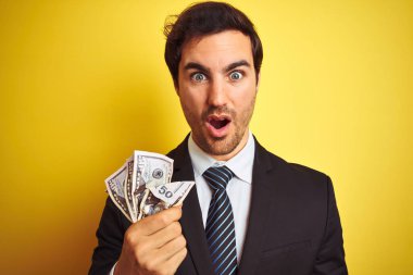 İzole sarı arka plan üzerinde dolar tutan takım elbise giyen genç yakışıklı işadamı sürpriz bir yüz ile şok korkmuş, korku ve korku ifade ile heyecanlı