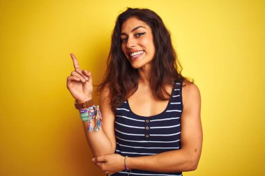 İzole edilmiş sarı arka planda duran çizgili tişört giyen genç ve güzel bir kadın yüzünde kocaman bir gülümsemeyle kameraya el ile işaret ediyor..