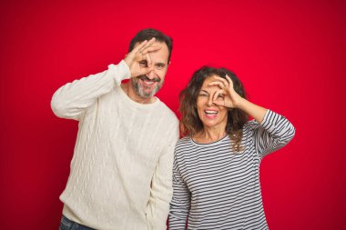 Güzel orta yaşlı bir çift, izole edilmiş kırmızı arka plan üzerine kışlık süveter giyip el hareketleriyle gülümsüyor ve mutlu yüzleriyle parmaklarının arasından bakıyor..
