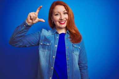 Genç, güzel, kızıl saçlı bir kadın mavi izole edilmiş arka plan üzerinde duruyor gülümsüyor ve el hareketleriyle küçük beden işareti yapıyor ve kameraya bakıyor. Ölçü kavramı.
