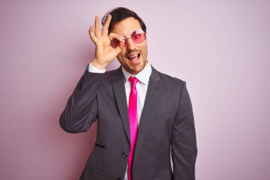 Genç yakışıklı iş adamı takım elbise ve güneş gözlüğü takıyor pembe arka planda el hareketleriyle gülümsüyor ve mutlu yüzle parmaklarının arasından bakıyor..