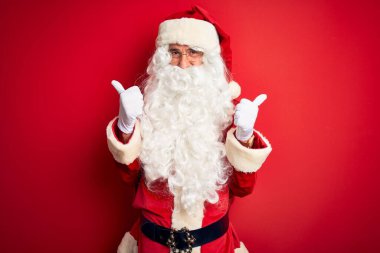 Noel Baba kostümü giymiş orta yaşlı yakışıklı bir adam izole edilmiş kırmızı arka plan işareti üzerinde duruyor. Elleriyle olumlu jest yapıyor, gülümsüyor ve mutlu. Neşeli ifade ve kazanan jest.