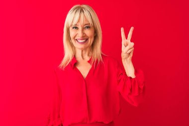 Soyutlanmış kırmızı arka plan üzerinde duran zarif gömlekli orta yaşlı bir kadın iki numaralı parmaklarıyla işaret ederken kendinden emin ve mutlu gülümsüyor..