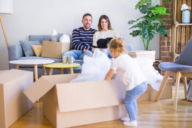 Güzel aile, anne-babalar kanepede kahve içme oturmuş karton kutular etrafında yeni evde oynayan çocuğuna bakıyor