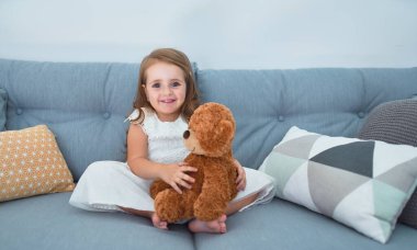 Sevimli sarışın bebek mutlu gülümsüyor. Kanepede oturup evde oyuncak ayıyla oynamak.