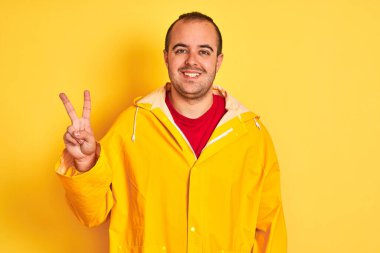 Yağmurluk giyen genç adam izole edilmiş sarı arka planda duruyor ve iki numaralı parmaklarıyla işaret ederken kendinden emin ve mutlu bir şekilde gülümsüyor..