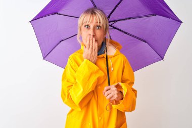 İzole edilmiş beyaz arka plan üzerine orta yaşlı kadın yağmurluk ve mor şemsiye giyiyor. Ağzı kapalı, hata yaptığı için utanmış, korku ifadesi, sessizlikten korkmak, gizli bir konsept.