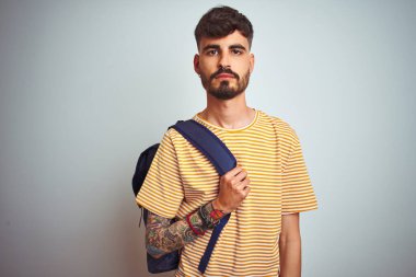 Sırt çantasında dövmesi olan genç bir öğrenci izole edilmiş beyaz bir altyapının başında duruyor. Zeki bir yüz ifadesi var ve ciddi düşünüyor.