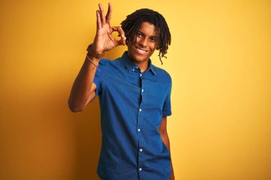 Rastgele kot gömlekli Afro adam izole edilmiş sarı arka planda dikiliyor, gülümsüyor ve el ve parmaklarıyla tamam işareti yapıyor. Başarılı ifade.