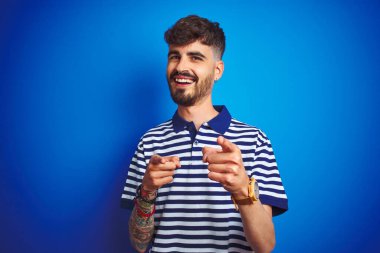 İzole edilmiş mavi arka planda duran çizgili polo giyen genç adam mutlu ve komik yüzlü parmaklarını kameraya doğrultuyor. İyi enerji ve titreşimler.