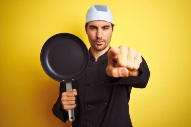 Üniformalı ve şapkalı genç aşçı, izole edilmiş sarı arka planda, kamerayı işaret ediyor. Ve sen, el işareti, ön taraftan olumlu ve kendinden emin bir hareket.