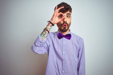 Mor gömlekli ve papyonlu genç adam izole edilmiş beyaz arka planda şaşırmış yüz ifadesiyle şaşırmış bir şekilde hareket ediyor. İnançsız ifade.