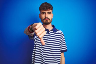 İzole edilmiş mavi arka planda duran çizgili polo giyen genç adam mutsuz ve kızgın görünüyor. Reddedilmiş ve olumsuz bir hareket yapıyor. Kötü ifade.