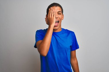 Mavi tişörtlü genç yakışıklı Arap adam izole edilmiş beyaz arka planda duruyor. Bir gözünü eliyle kapatıyor. Yüzünde kendine güvenen bir gülümseme ve sürpriz bir duygu var..