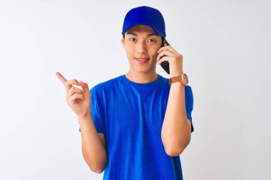 Şapkalı Çinli teslimatçı, izole edilmiş beyaz arka planda akıllı telefondan konuşuyor. Elleri ve parmakları kenarı işaret ederek çok mutlu.