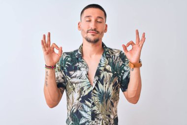 İzole edilmiş arka planda Hawaii tişörtü giyen genç yakışıklı adam rahatla ve parmaklarıyla meditasyon hareketi yaparken gözleri kapalı gülümse. Yoga konsepti.
