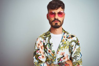 Tatilde dövmeli, güneş gözlüğü takan bir adam izole edilmiş beyaz arka plan şüphecisi ve gergin, yüzünde çapraz kollu tasvip etmeyen bir ifade var. Negatif kişi.