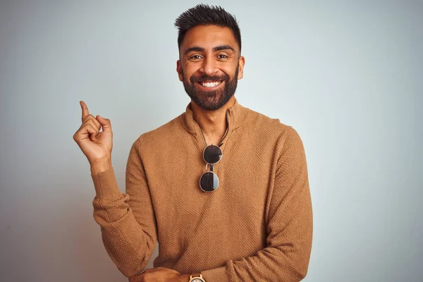 Indian man wearing elegant sweater and sunglasses standing over isolated white background with a big smile on face, pointing with hand and finger to the side looking at the camera.