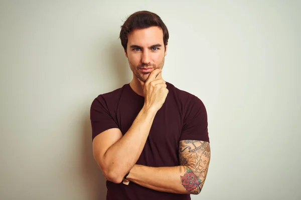 Young handsome man with tattoo wearing purple casual t-shirt over isolated white background looking confident at the camera with smile with crossed arms and hand raised on chin. Thinking positive.