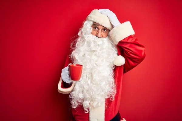 Senior man wearing Santa Claus costume holding cup of coffee over isolated red background stressed with hand on head, shocked with shame and surprise face, angry and frustrated. Fear and upset for mistake.